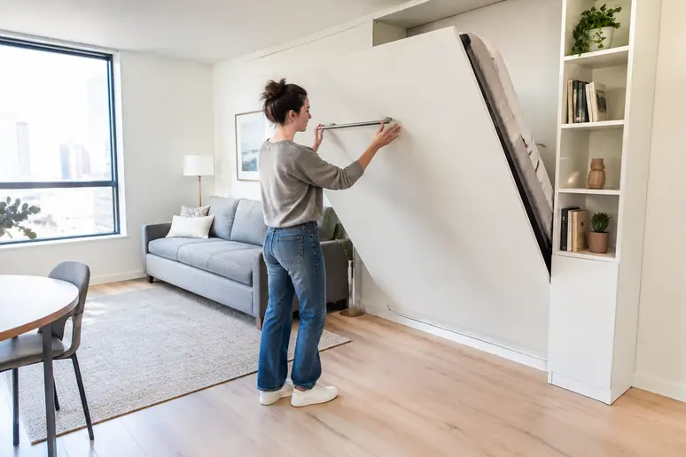 Personne repliant un lit mural rabattable dans un studio lumineux pour libérer de l'espace au sol.